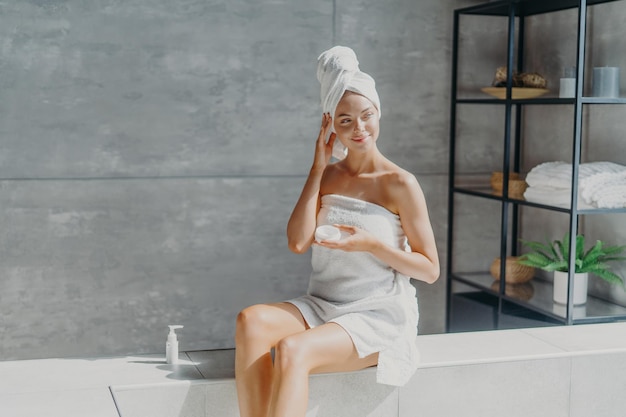 Health beauty and pampering concept Beautiful dreamy young Eurpean woman looks away thoughtfully applies body cream sits in bathroom wrapped in white soft towel uses nice cosmetic product