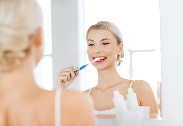 health care, dental hygiene, people and beauty concept - smiling young woman with toothbrush cleaning teeth and looking to mirror at home bathroom