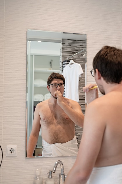 Health care , dental hygiene. Young adult man brushing teeth in bathroom, looking at the mirror