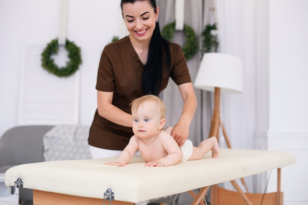 Health care and medical concept. Happy baby having massage with professional female masseuse. Kind and friendly children's doctor. Children's massage on the couch in a modern cozy room.