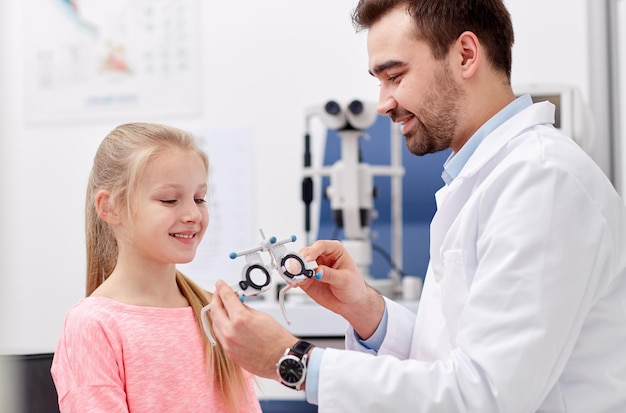 health care, medicine, people, eyesight and technology concept - optometrist with trial frame checking girl patient vision at eye clinic or optics store