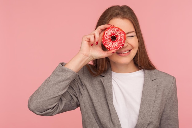 Health care and nutrition. Portrait of cheerful businesswoman in suit jacket looking through sweet doughnut, having fun with pastry, peeking into donut and smiling. indoor studio shot, pink background