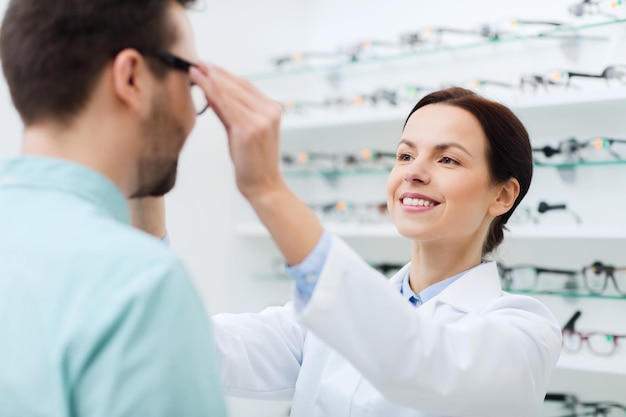 health care, people, eyesight and vision concept - female optician putting on glasses to man at optics store