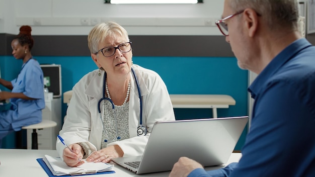 Health specialist taking notes at checkup examination in cabinet, having discussion with senior patient at appointment. Doctor writing health care report on documents to cure disease.