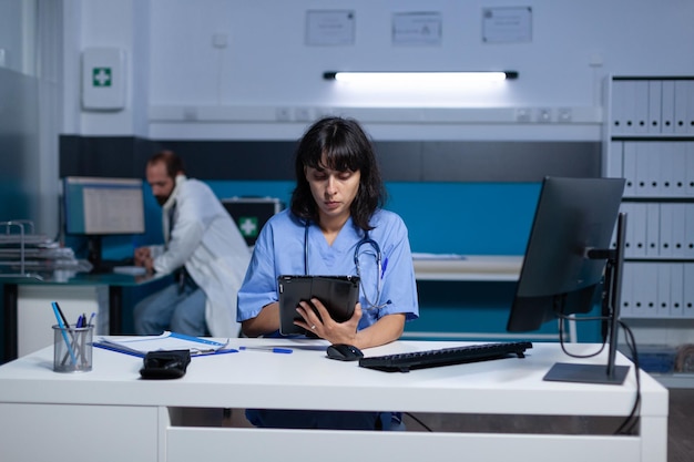 Healthcare assistant working on digital tablet for checkup appointment and treatment overtime. Medical specialist looking at device screen and computer. Nurse with expertise working late