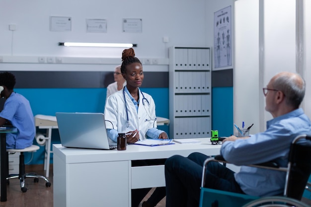 Healthcare clinic medic expert conversating with sick senior patient in wheelchair about illness symptoms and treatment schedule. Hospital doctor talking with elder person with disability about recove