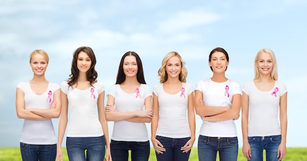 healthcare, people and medicine concept - group of smiling women in blank t-shirts with pink breast cancer awareness ribbons blue sky and grass background