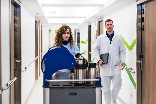 Healthcare staff in blue pushing a coffee cart making eye contact with the camera with a doctor