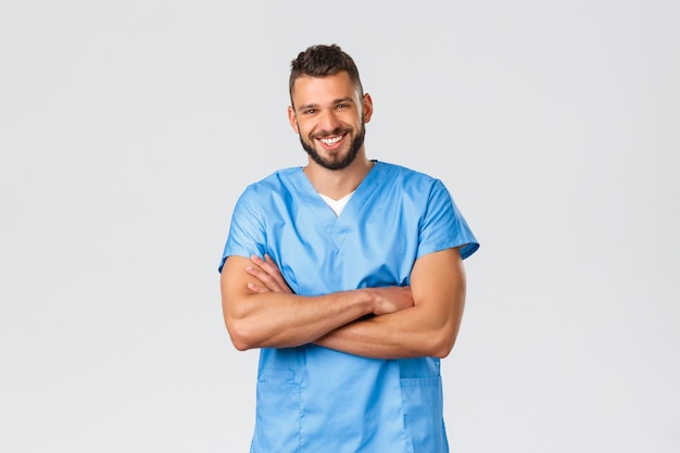 Healthcare workers, medicine, covid-19 and pandemic self-quarantine concept. Smiling handsome, hispanic doctor, male nurse in blue scrubs, cross arms chest, laughing camera cheerful.