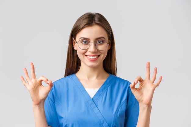 Healthcare workers, medicine, insurance and covid-19 pandemic concept. Confident and optimistic female nurse, doctor in blue scrubs and glasses, show okay sign, guarantee best service quality