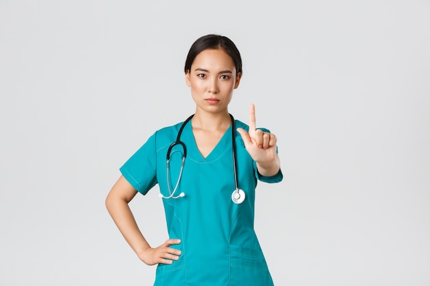 , healthcare workers, pandemic concept. Serious-looking professional female asian doctor in scrubs, physician shaking finger in prohibition, warning gesture, forbid something