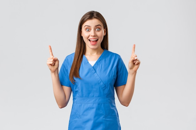 Healthcare workers, prevent virus, insurance and medicine concept. Excited, happy nurse or doctor in blue scrubs smiling from good news, pointing fingers up, show patients great news