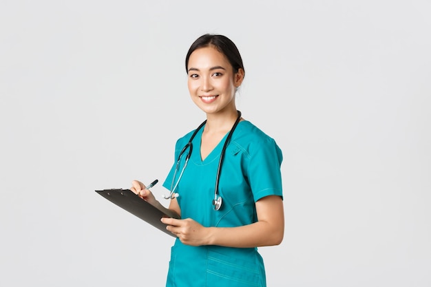 Healthcare workers preventing virus quarantine campaign concept Smiling friendly female doctor nurse in scrubs writing dorn results analysis at clipboard standing white background