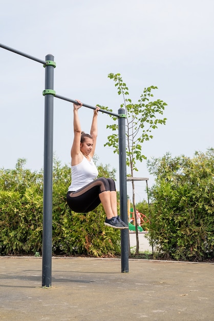 Healthy and active lifestyle. Sports and fitness. Happy woman in white t shirt working out on the sports ground in sunny summer day, training her abs on the bars