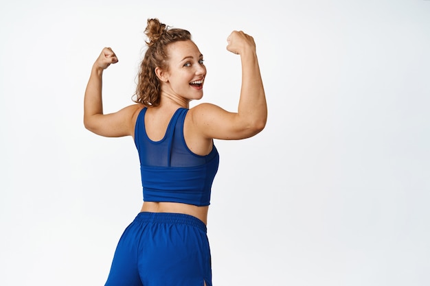 Healthy and active sportswoman showing muscles, turn behind and laughing, flexing biceps on arms, feeling strong after workout, standing on white.