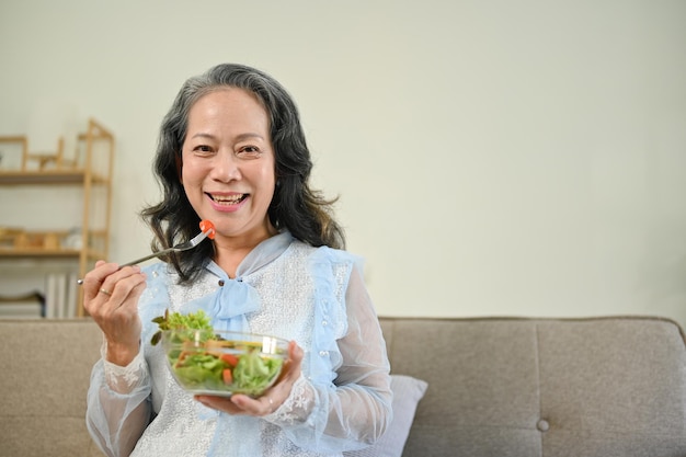 Healthy Asian 60s aged woman enjoys eating tasty Buddha salad mix bowl in her living room