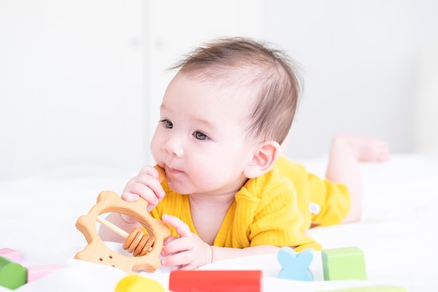 healthy asian baby girl in yellow bodysuit plays with wooden toy teether for teeth on white bedding