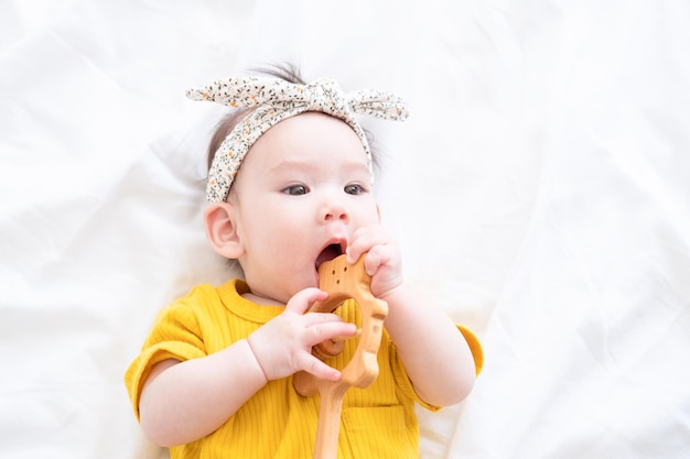 healthy asian baby girl in yellow bodysuit plays with wooden toy teether for teeth on white bedding