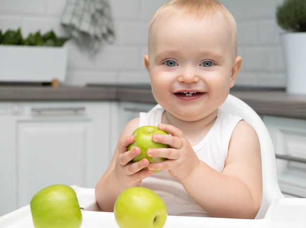 Healthy baby boy big blu eyes sitting in a child's chair in the kitchen eats green apples.