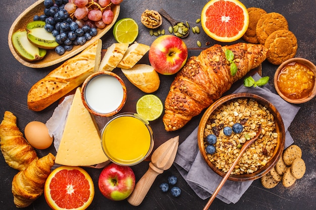 Healthy balanced breakfast on a dark background. Muesli, milk, juice, croissants, cheese, biscuits.