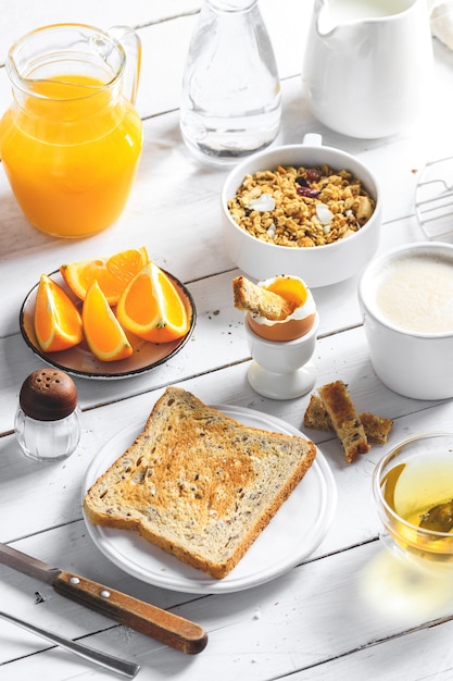 Healthy breakfast eating concept, various morning food - pancakes, soft-boiled egg, toast, oatmeal, granola, fruit, coffee, tea, orange juice, milk on white wooden table