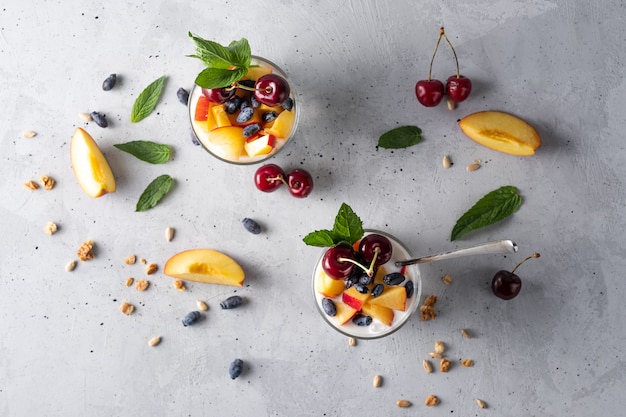 Healthy breakfast in a glass with fresh fruits: pomegranate, cherry, nectarines, honeysuckle, yogurt and granola on a grey background. Shallow depth of field with selective focus