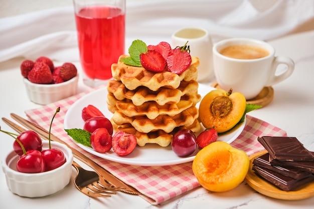 Healthy Breakfast set with Belgian waffles with strawberries, apricots, cherries, juice and a cup of black coffee and bitter chocolate on white stone table background