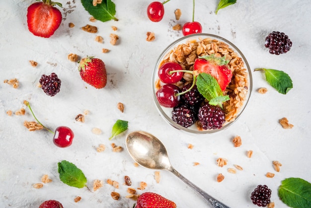 Healthy breakfast. Summer berries and fruits. Homemade Greek yoghurt with granola, blackberries, strawberries, cherries and mint. On white concrete stone table, in glasses. 