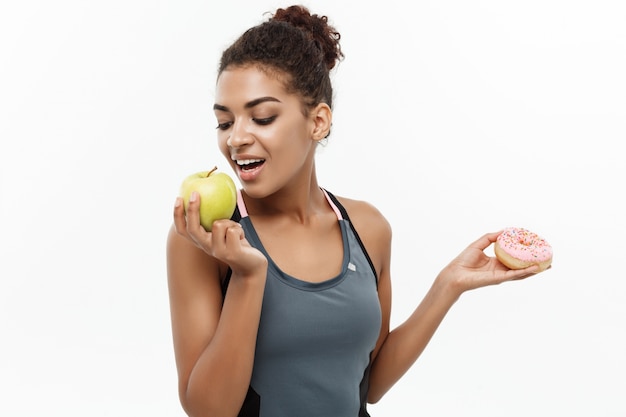 Healthy and diet concept - Beautiful sporty African American make a decision between donut and green apple. Isolated on white background.