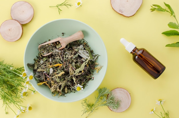 Healthy dry herbs in natural medicine bowl and water extract in bottle on yellow paper background