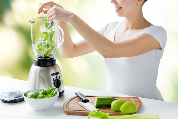 healthy eating, cooking, vegetarian food, dieting and people concept - close up of young woman with blender and green vegetables making detox shake or smoothie over natural background