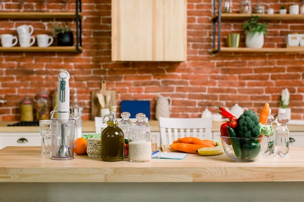 Healthy eating and diet concept - natural food on table. Vegetables, fruits, rice, olive oil, glasses, blender on the wooden table in light modern kitchen