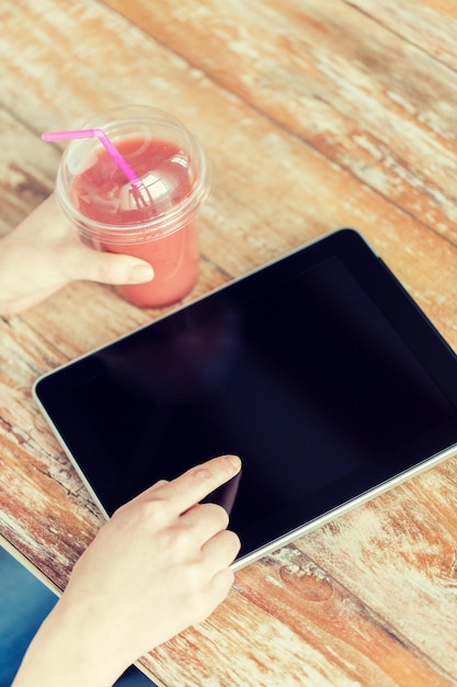 healthy eating, diet, technology and people concept - close up of woman hands with cup of smoothie pointing finger to tablet pc computer black blank screen on wooden table