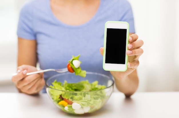 healthy eating, dieting, technology, food and people concept - close up of young woman with smartphone eating vegetable salad at home