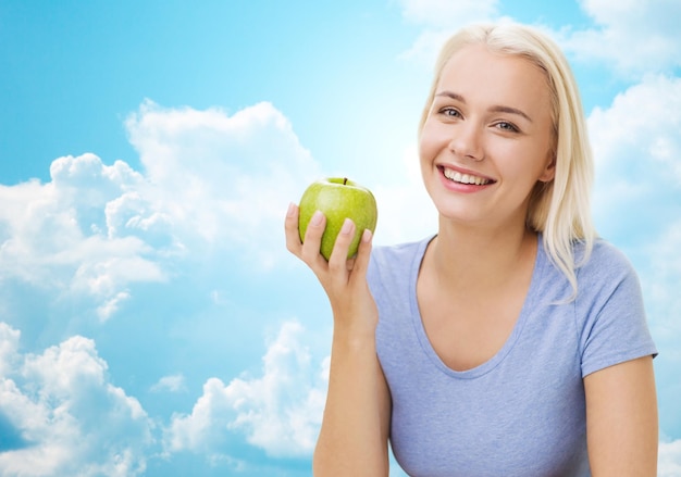 healthy eating, organic food, fruits, diet and people concept - happy woman eating green apple over blue sky and clouds background