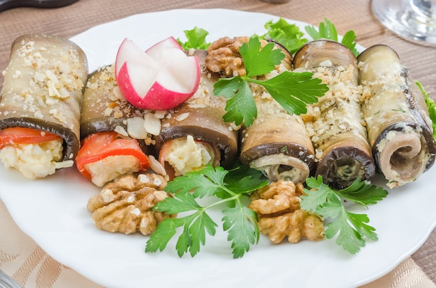 Healthy eggplant rolls stuffed with cheese, served with vegetables and herbs close-up on a white plate on a dark wooden background