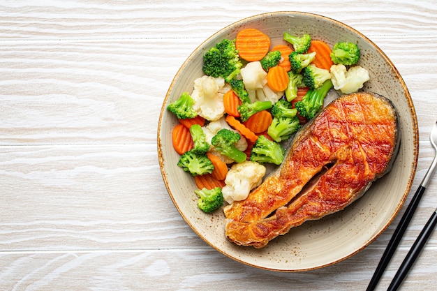 Healthy fish dinner: grilled salmon fish steak with vegetables salad on ceramic plate with fork and knife on white wooden background from above with copy space, clean and diet eating