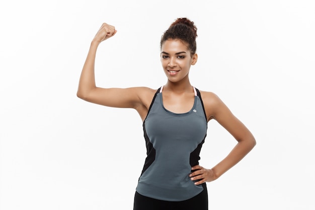 Healthy and Fitness concept - Portrait of young beautiful African American showing her strong muscle with confident cheerful facial expression. Isolated on white studio background.