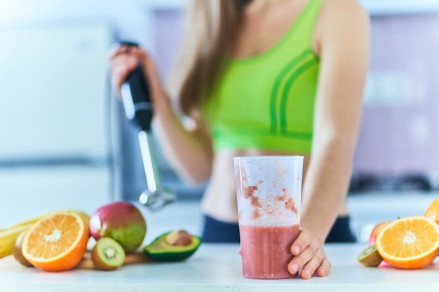 Healthy fitness woman in sportswear prepares a fresh fruit smoothie using a hand blender at home in the kitchen.