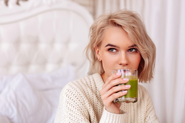 Healthy green juice. Close up of blonde-haired young woman drinking healthy green juice in bedroom