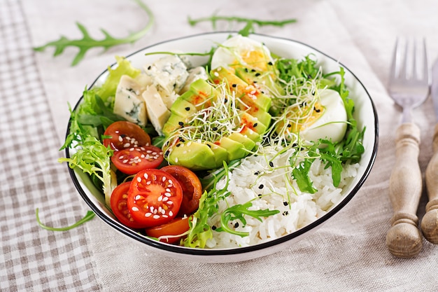 Healthy green vegetarian buddha bowl lunch with eggs, rice, tomato, avocado and blue cheese on table. 