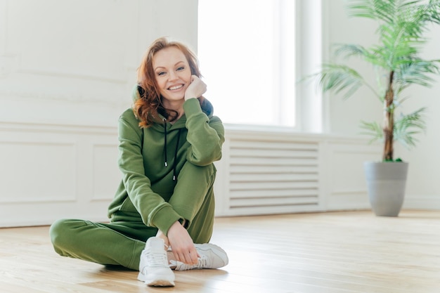 Healthy lifestyle concept Beautiful slim young woman with red wavy hair keeps hand under chin smiles happily dressed in sportswear has flexible body sits on floor Female has home workout