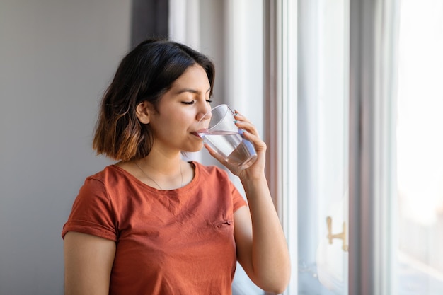 Healthy liquid arab woman drinking water from glass while standing near window