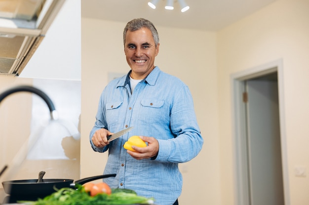 Healthy lunch time! Adult cheerful man in blue shirt standing near stove, holding yellow paper and knife, looking at camera, smiling. Vegan food concept. Man adding some vegetables to the dish.