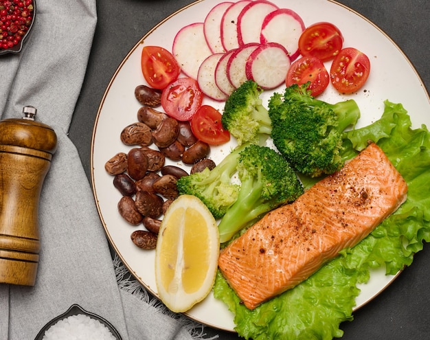 Healthy lunch with grilled salmon on green lettuce next to vegetables tomatoes radishes broccoli and a portion of beans