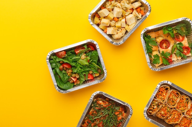 Healthy meals delivery. Casserole, vegetarian rolls, fresh and baked vegetables salads in foil take away boxes on yellow background. Eating right concept, copy space, top view.