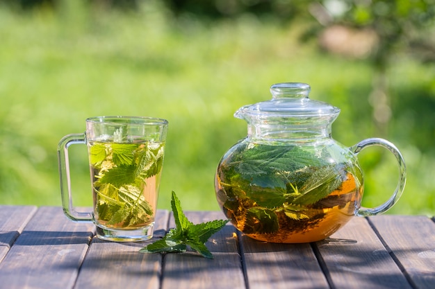 Photo healthy nettle tea in a glass tea pot and mug in the summer garden on wooden table. close up herbal tea from the green petals of nettle on nature background