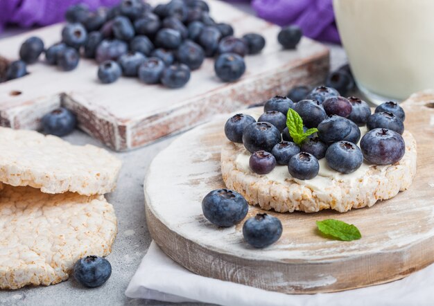 Healthy organic rice cakes with ricotta and fresh blueberries on wooden board and glass of milk