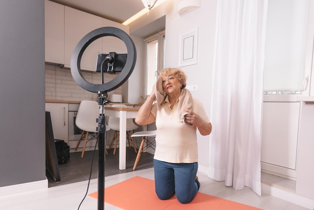 A healthy pensioner sits on the bedroom floor in front of a tripod for a camera, shoots a fitness video tutorial for the elderly.