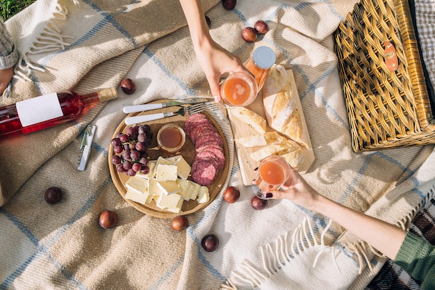 A healthy picnic on summer holidays with freshly baked bread, cheese and salami is laid out on a brown plaid. Girls drink multifruit juice in the park.